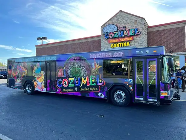 A fully wrapped bus for Mi Cozumel Mexican restaurant. The bus is purple, with vibrant neon colored lettering and photos of their signature margaritas and flaming fajitas. This design shows that restaurant advertising with public transit can be fun and dynamic!