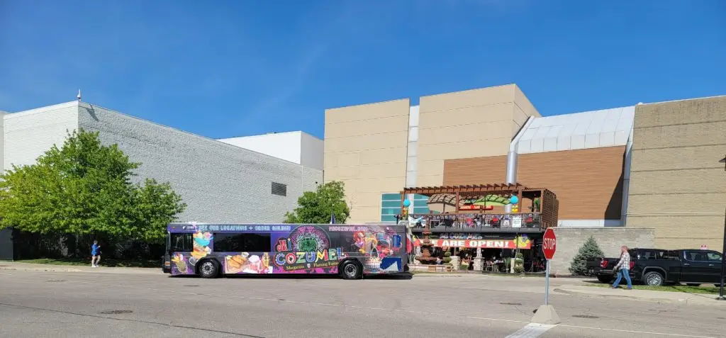 Mi Cozumel's full wrap bus sitting in front of one of their locations. The bus is purple with images of their food options and drink options contrasting with the background. Their logo is in the center. People are walking towards the restaurant and looking at the bus.