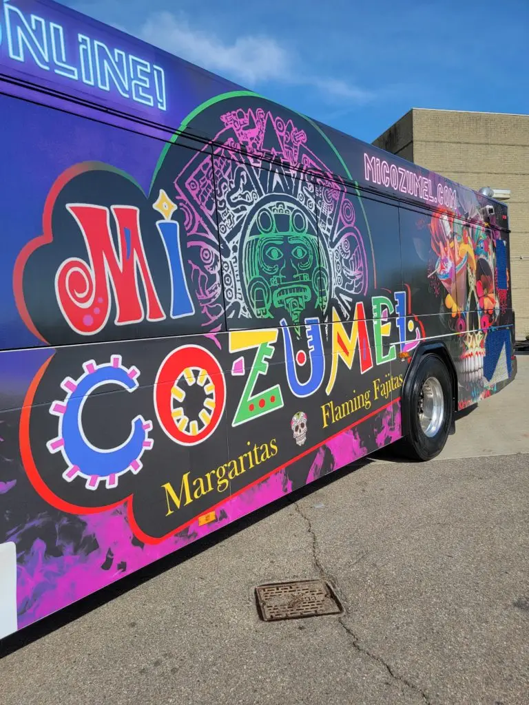 A close up shot of the Mi Cozumel bus. The logo is in full view, it's primary colors popping off the pink and purple background. There is a skull behind the logo.