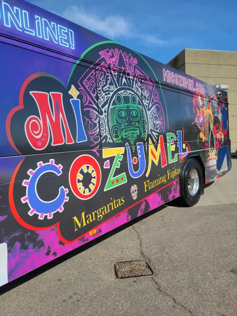 A close up shot of the Mi Cozumel bus. The logo is in full view, it's primary colors popping off the pink and purple background. There is a skull behind the logo.