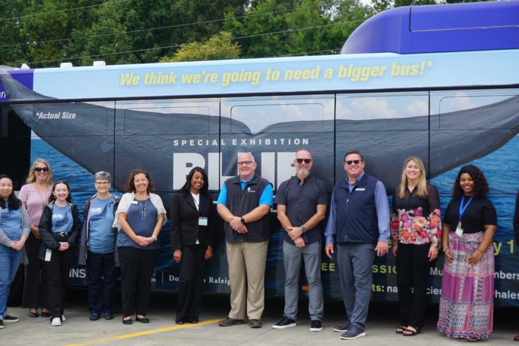 Group photo from the GoDurham launch event. Features a group of people standing and smiling in front of a city bus with a large advertisement on it that has a blue whale tail, advertising for the Museum's new exhibition.